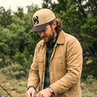 A man wearing a tan crewneck t-shirt and a tan 3 Up 3 Down baseball hat, fixing his car in a rustic wooden setting.