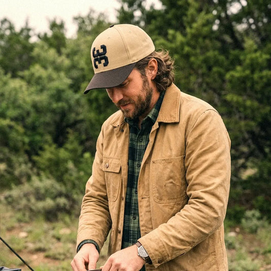 A man wearing a tan crewneck t-shirt and a tan 3 Up 3 Down baseball hat, fixing his car in a rustic wooden setting.