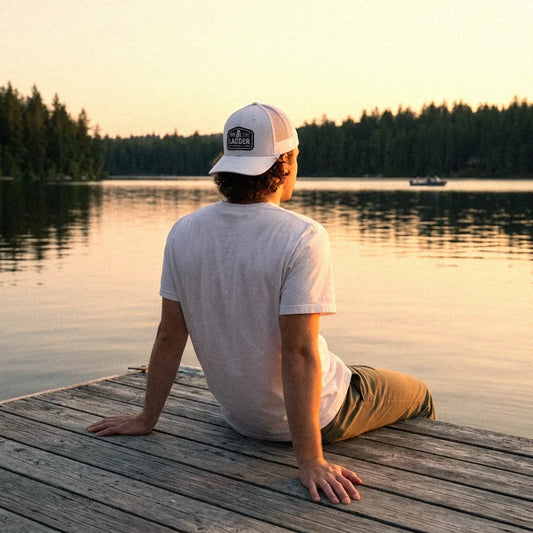 A man sits on a wooden dock by a calm lake at sunset, seen from behind. He is wearing a Climb the Ladder white trucker hat, a white t-shirt, and khaki pants.