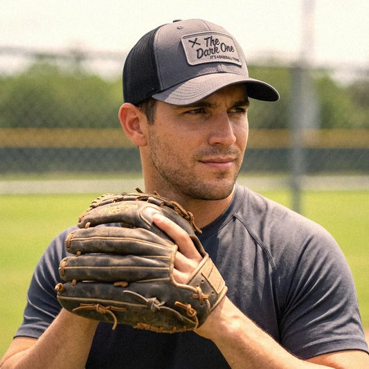 A man on a baseball field wearing a The Dark One low profile trucker hat in charcoal, a charcoal athletic t-shirt, and holding a brown leather baseball glove.