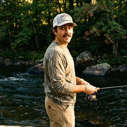 A man standing by a flowing river in a wooded outdoor setting, wearing a camouflage long-sleeve shirt, khaki pants, and a white Died A Hero trucker hat, while holding a fishing rod.