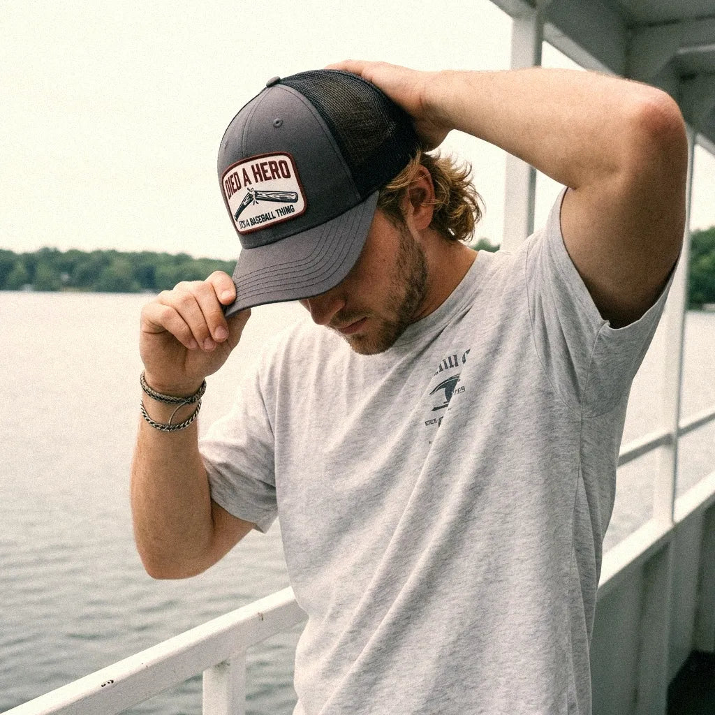 A man wearing a charcoal and black Died A Hero trucker hat on, standing on a boat deck with water in the background