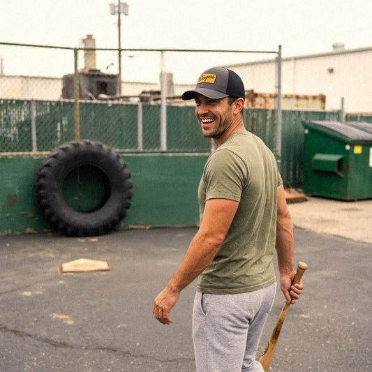 A man wearing an olive green t-shirt, gray sweatpants, and a charcoal Dingers baseball cap stands in an outdoor industrial yard holding a baseball bat.