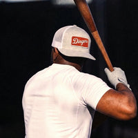 A man from behind wearing a white t-shirt and Dingers snapback trucker hat in heather gray, holding a wooden baseball bat against a dark background.