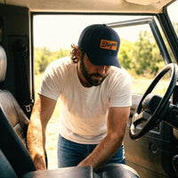 A man with a beard wearing a white t-shirt, blue jeans, and a navy blue Dingers trucker hat snapback leans over the passenger side of a vehicle.