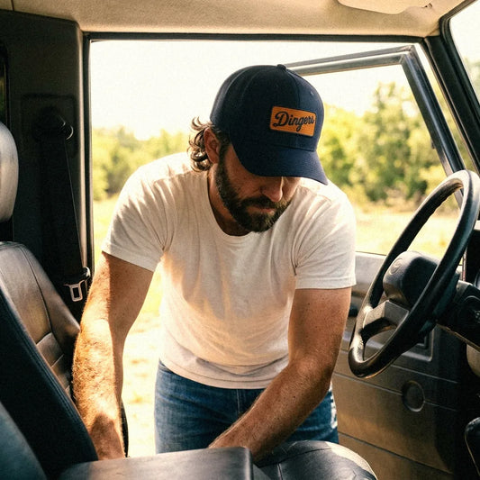 A man with a beard wearing a white t-shirt, blue jeans, and a navy blue Dingers trucker hat snapback leans over the passenger side of a vehicle.