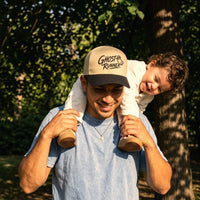 A man outdoors carrying a child on his shoulders, wearing a Ghost Runner snapback hat in khaki and black. The background is a sunlit, leafy park setting.