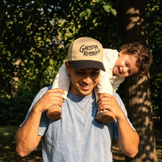 A man outdoors carrying a child on his shoulders, wearing a Ghost Runner snapback hat in khaki and black. The background is a sunlit, leafy park setting.