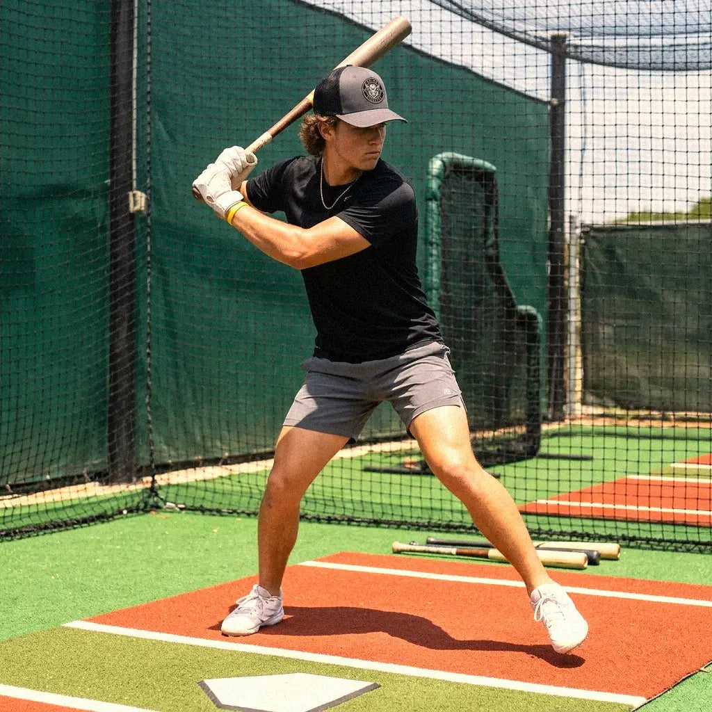 A man in a black t-shirt, gray shorts, and a charcoal Going Yicketty hat swings a wooden baseball bat on a batting practice field with a green fence and red hitting mat.