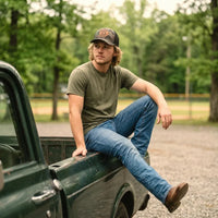 A man with blonde hair wearing a green t-shirt, blue jeans, and a camouflage Hunting Fastballs baseball cap sits casually on the bed of a dark green pickup truck in an outdoor wooded setting.