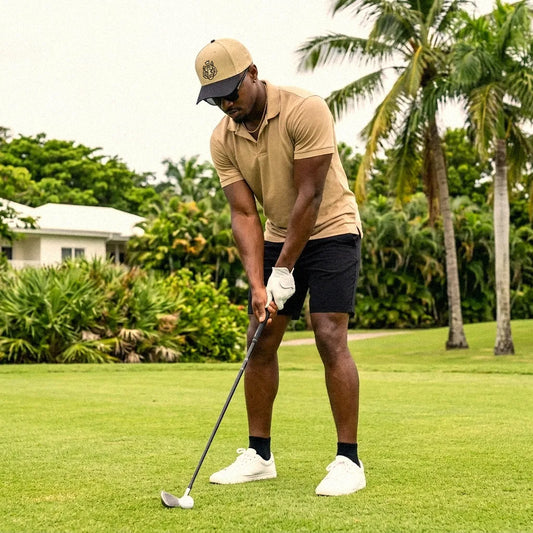 A man wearing a tan polo shirt, black shorts, white golf shoes, and a tan NOBLETIGER snapback hat is shown mid-swing on a golf course with palm trees and green grass in the background.