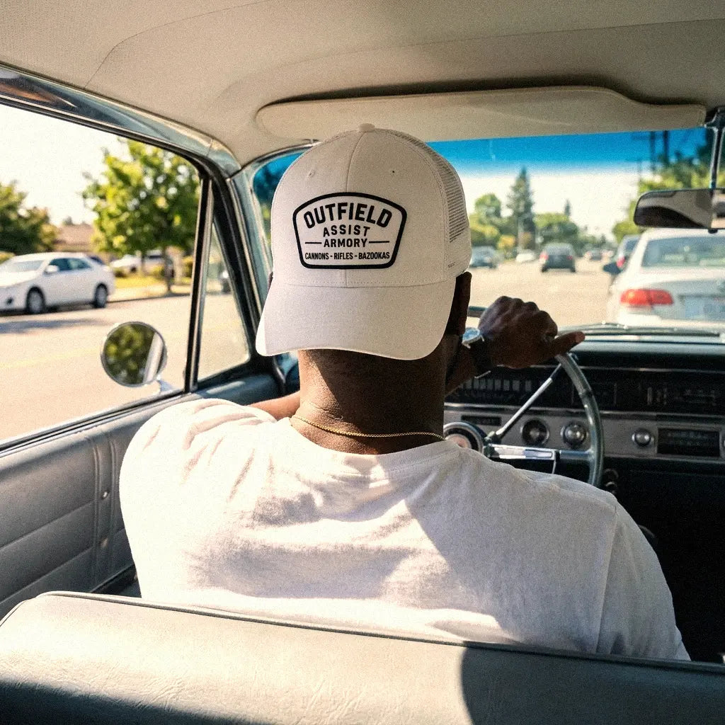 A man driving a vintage car, viewed from behind, wearing a white Outfield Assist Armory low profile trucker hat.