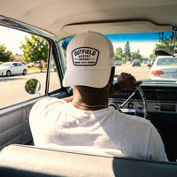 A man driving a vintage car, viewed from behind, wearing a white Outfield Assist Armory low profile trucker hat.