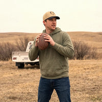 A man wearing an olive green hoodie, blue jeans, and a tan Paint the Corners snapback hat stands in a dry grass field holding a football, with a white pickup truck blurred in the background.