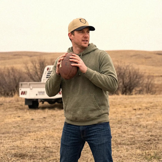 A man wearing an olive green hoodie, blue jeans, and a tan Paint the Corners snapback hat stands in a dry grass field holding a football, with a white pickup truck blurred in the background.