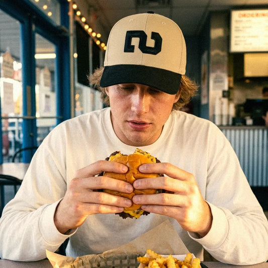 A man wearing a khaki and black Turn Two 6 panel snapback hat, wearing a white long-sleeve shirt, and holding a burger in a restaurant setting.