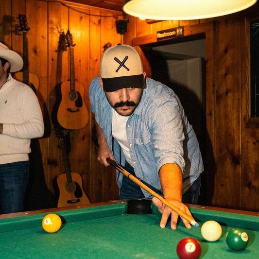 Man wearing jeans, a white undershirt, and a blue button up shirt with a Twin Bills 6 panel snapback hat in khaki with a black bill leaning over a pool table in a bar