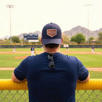 A man viewed from behind at a baseball game, wearing a navy blue t-shirt and a navy blue Twin Bills trucker hat. He is leaning on a yellow fence in front of a baseball field with mountains in the background.