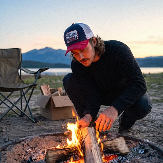 A man wearing an Uncle Charlie trucker hat and a black sweater crouches by a campfire outdoors at sunset, with a folding chair and cardboard box in the background near mountains and water.